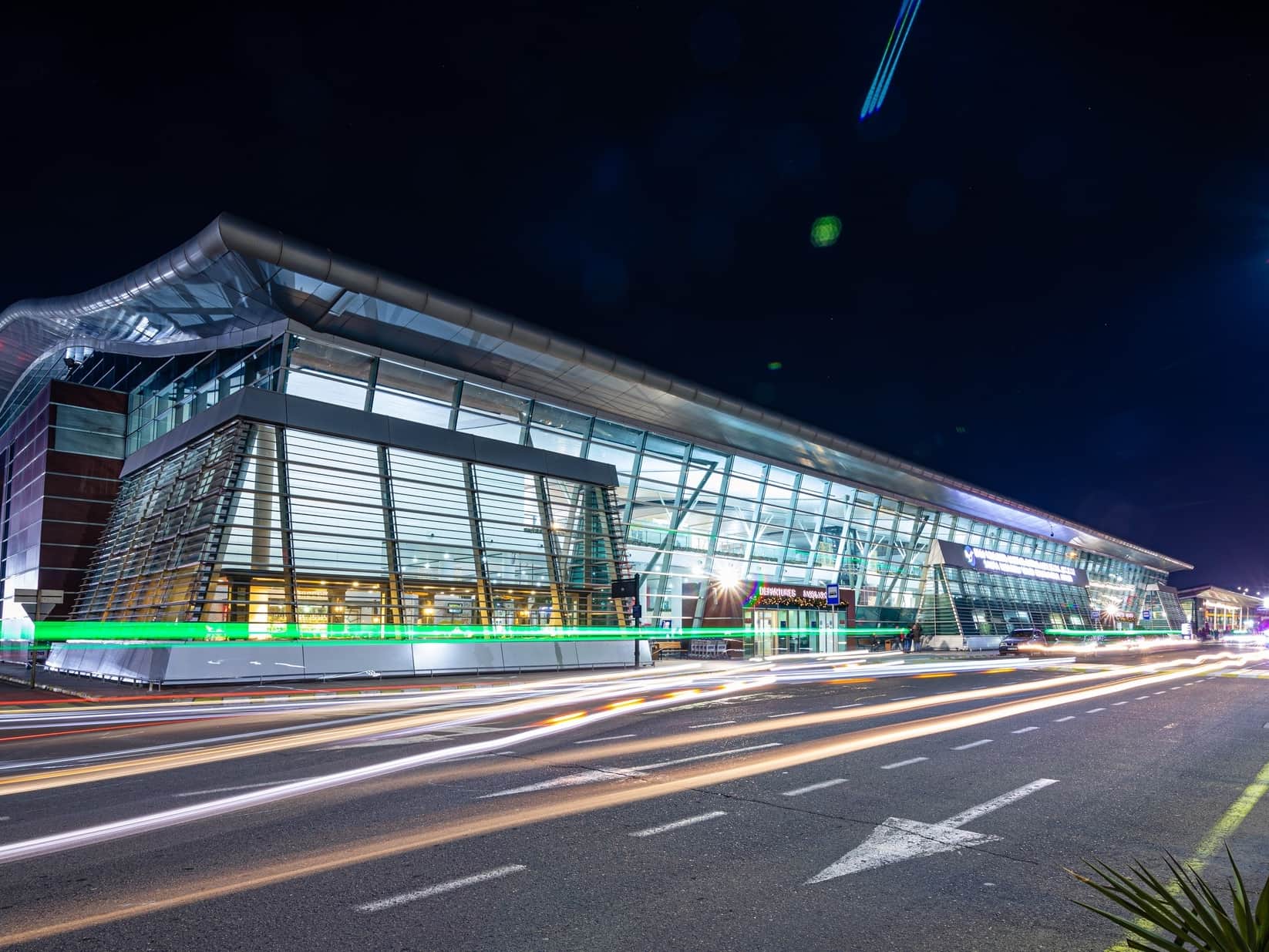 The illuminated terminal building at Tbilisi International Airport at night, with traffic passing the departures ramp